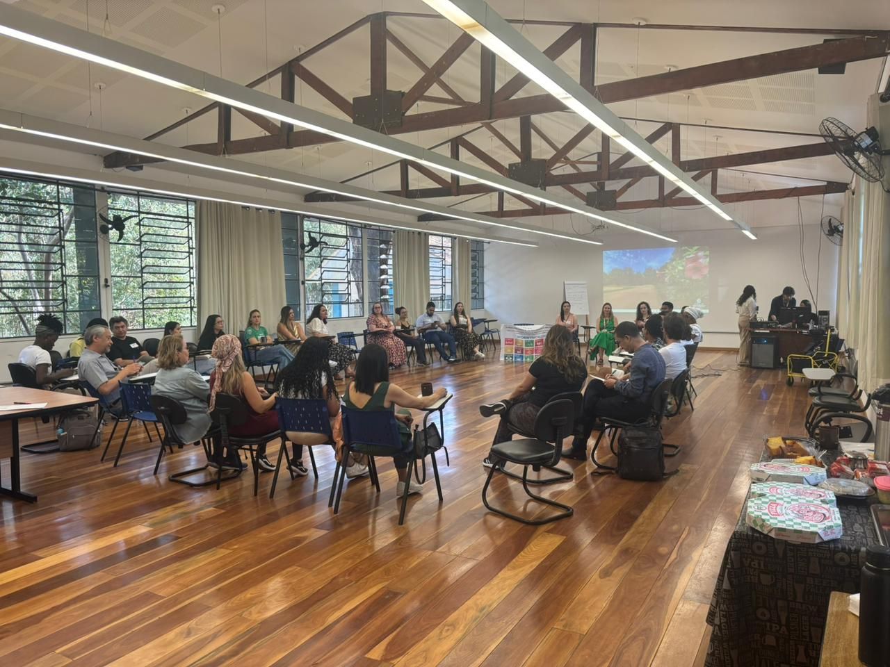 Sala ampla com piso de madeira, onde um grupo de pessoas está sentado em cadeiras organizadas em círculo para uma atividade ou roda de conversa. Ao fundo, há uma projeção na parede e duas pessoas próximas a equipamentos de som e computador. À direita, sobre uma mesa, há alimentos e bebidas organizados.