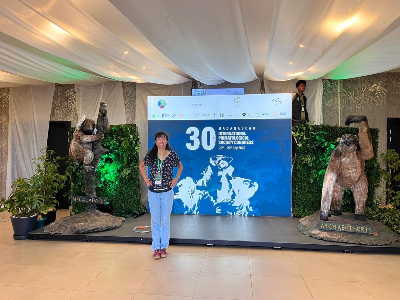 Uma fotografia de uma mulher em pé na frente de um palco com um banner azul. O banner tem o logo do "30th International Primatological Society Congress" e as datas "20-25th July 2025". O palco está decorado com duas estátuas de primatas extintos, uma com a etiqueta "MEGALADAPIS" e a outra "ARCHAEOINDRIS". Há algumas plantas e um homem em pé ao fundo.