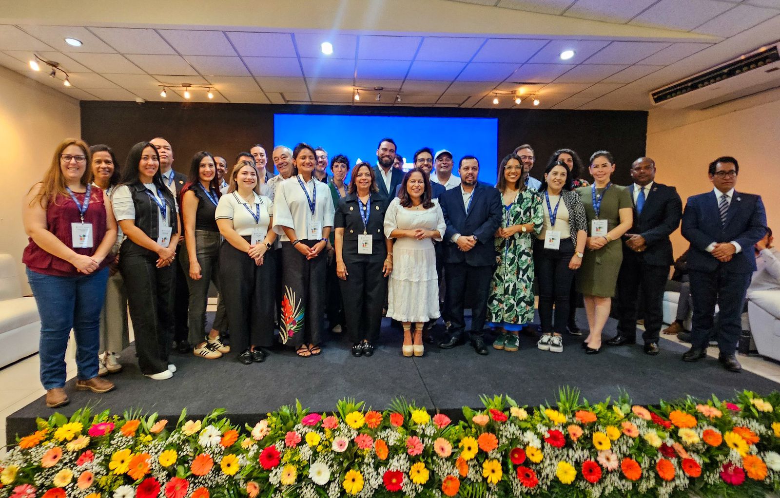 A imagem mostra um grupo de cerca de 26 pessoas posando para uma foto coletiva sobre um palco. Ao fundo, há uma tela grande com fundo azul e iluminação quente no teto. As pessoas vestem roupas formais ou sociais, predominando tons neutros como preto, branco, azul-marinho e verde, além de alguns padrões estampados.  Na parte inferior da imagem, à frente do palco, há um arranjo extenso e vibrante de flores — nas cores amarelo, laranja, vermelho, rosa e branco — intercaladas com folhagem verde. O cenário transmite um clima de evento oficial ou congresso, com credenciais penduradas no pescoço de quase todos os participantes.