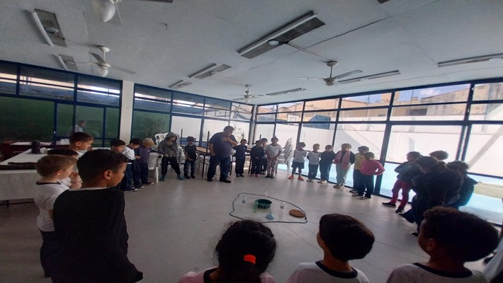 A primeira imagem mostra um grupo de crianças em um ambiente interno claro, com paredes de vidro azul-marinho e grandes janelas, formando um círculo em volta de um instrutor no centro. No chão, há um desenho no formato de um percurso, com objetos como baldes e tábuas. As crianças usam roupas casuais, muitas com camisetas brancas de gola preta.