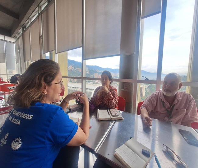 A segunda mostra uma reunião em uma sala envidraçada com vista para a cidade e montanhas ao fundo. Três pessoas estão em discussão, com cadernos e anotações sobre a mesa.