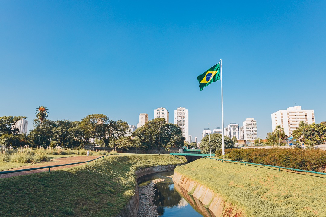 A imagem mostra um parque urbano com uma bandeira do Brasil hasteada em um mastro alto ao centro-direita. Em primeiro plano, há um pequeno canal de água cercado por gramado e caminhos para pedestres. Ao fundo, veem-se árvores e edifícios altos, indicando uma área urbana densamente povoada. O céu está limpo e azul, sugerindo um dia ensolarado.