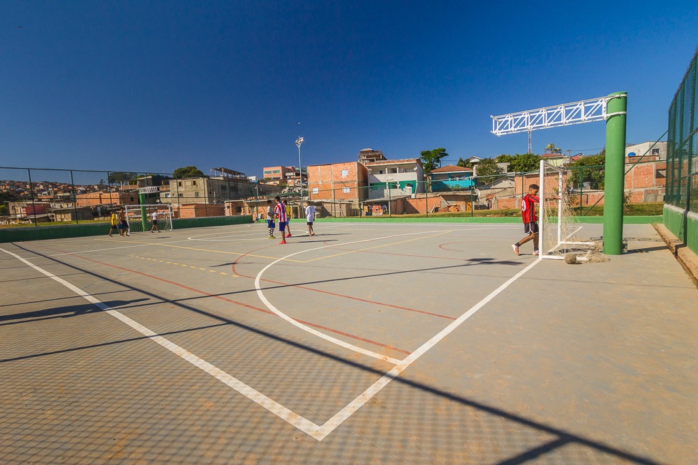 Crianças jogam futebol em uma quadra ao ar livre, rodeada por casas de tijolo e céu azul claro, promovendo lazer e inclusão social na comunidade.