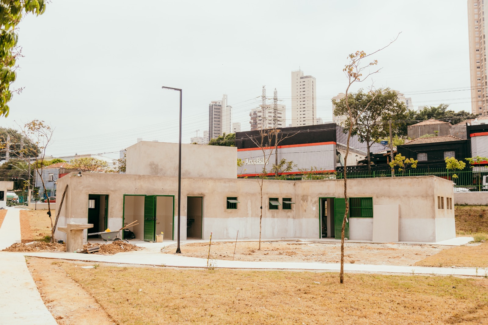 A imagem mostra um prédio em fase final de construção dentro de uma área aberta. A edificação tem formato retangular, paredes em tons de bege e cinza claro, e portas e janelas de cor verde. Algumas portas estão abertas, revelando o interior ainda em acabamento. Em frente ao prédio, há um canteiro de obras com restos de materiais, terra revirada e pequenas árvores recém-plantadas, de troncos finos e poucas folhas. O solo ao redor é de grama seca em tom amarelado, contrastando com a calçada de concreto clara que contorna o espaço. Ao fundo, aparecem casas baixas, um muro pintado de preto, cinza e vermelho, e prédios altos em tons de branco e bege, compondo a paisagem urbana. O céu está nublado em cinza claro, sem sol aparente.