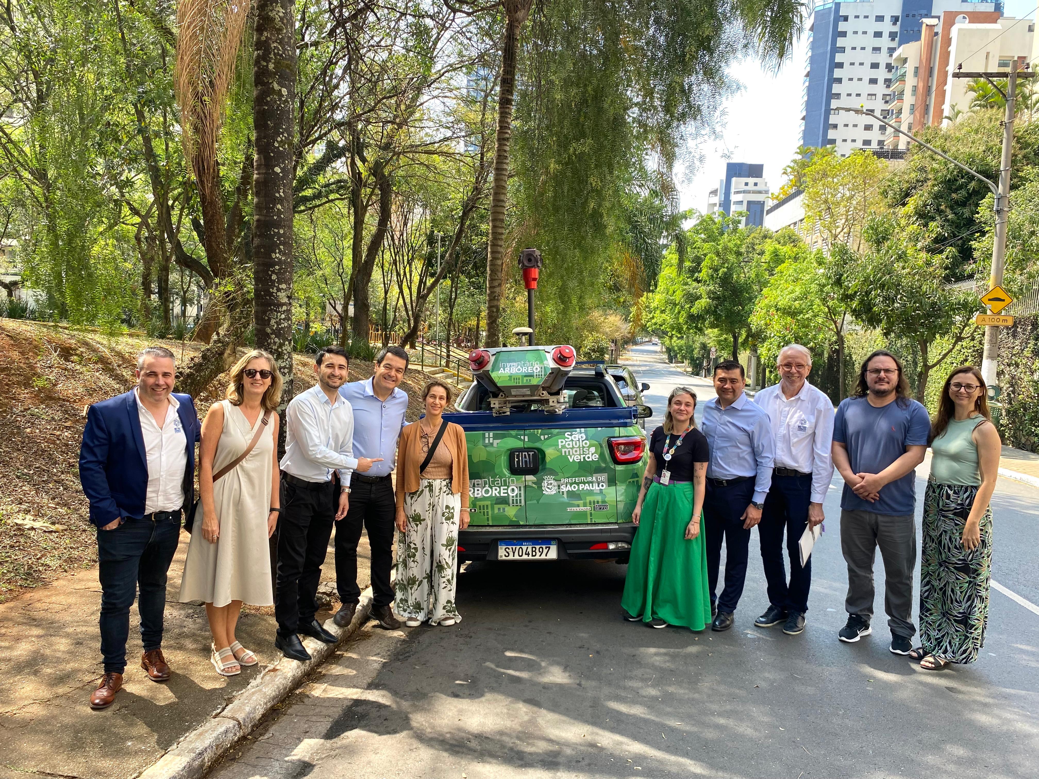 A imagem mostra um grupo de 11 pessoas posando para a foto em via pública arborizada, ao lado de um veículo verde identificado com logotipos da Prefeitura de São Paulo e do “Inventário Arbóreo”. O carro tem equipamentos de mapeamento instalados no teto, incluindo câmeras e sensores, além de adesivos com os dizeres “São Paulo mais verde” e “Inventário Arbóreo”. O grupo é formado por homens e mulheres, alguns em trajes sociais (camisa, calça social, blazer), outros em roupas mais casuais, como saias, vestidos e camisetas. Entre eles, destaca-se à esquerda a mulher de calça florida, que é a coordenadora da Divisão de Arborização Urbana, e à direita do carro, de calça social e camisa azul-clara, o secretário da SVMA, Rodrigo Ashiuchi. Ao fundo, aparecem árvores altas e prédios residenciais de médio e grande porte, compondo a paisagem urbana. O clima é de dia ensolarado, com céu parcialmente claro.