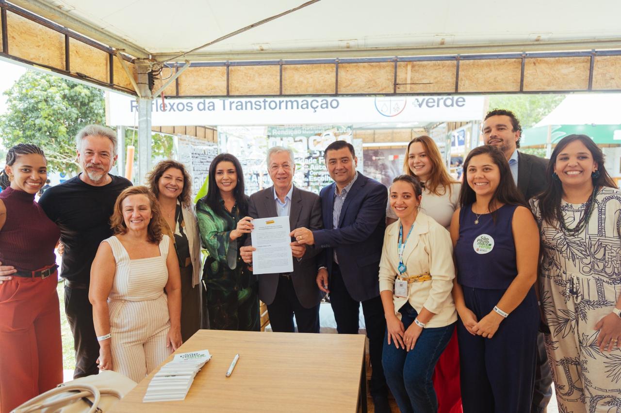 Um grupo de onze pessoas posa sorridente para uma foto embaixo de uma tenda clara. No centro, dois homens — um de terno cinza e outro de terno azul-marinho — seguram um documento oficial. À frente deles há uma mesa de madeira com uma caneta e alguns folhetos brancos empilhados.  As pessoas vestem roupas em tons variados: vinho, bege, verde, preto, branco, azul e estampado em azul e branco. Ao fundo, há uma faixa com as palavras “Reflexos da Transformação” e parte da palavra “Verde”. O ambiente é iluminado pela luz natural, com árvores e outras estruturas visíveis do lado de fora da tenda. A atmosfera é de celebração e formalidade leve.