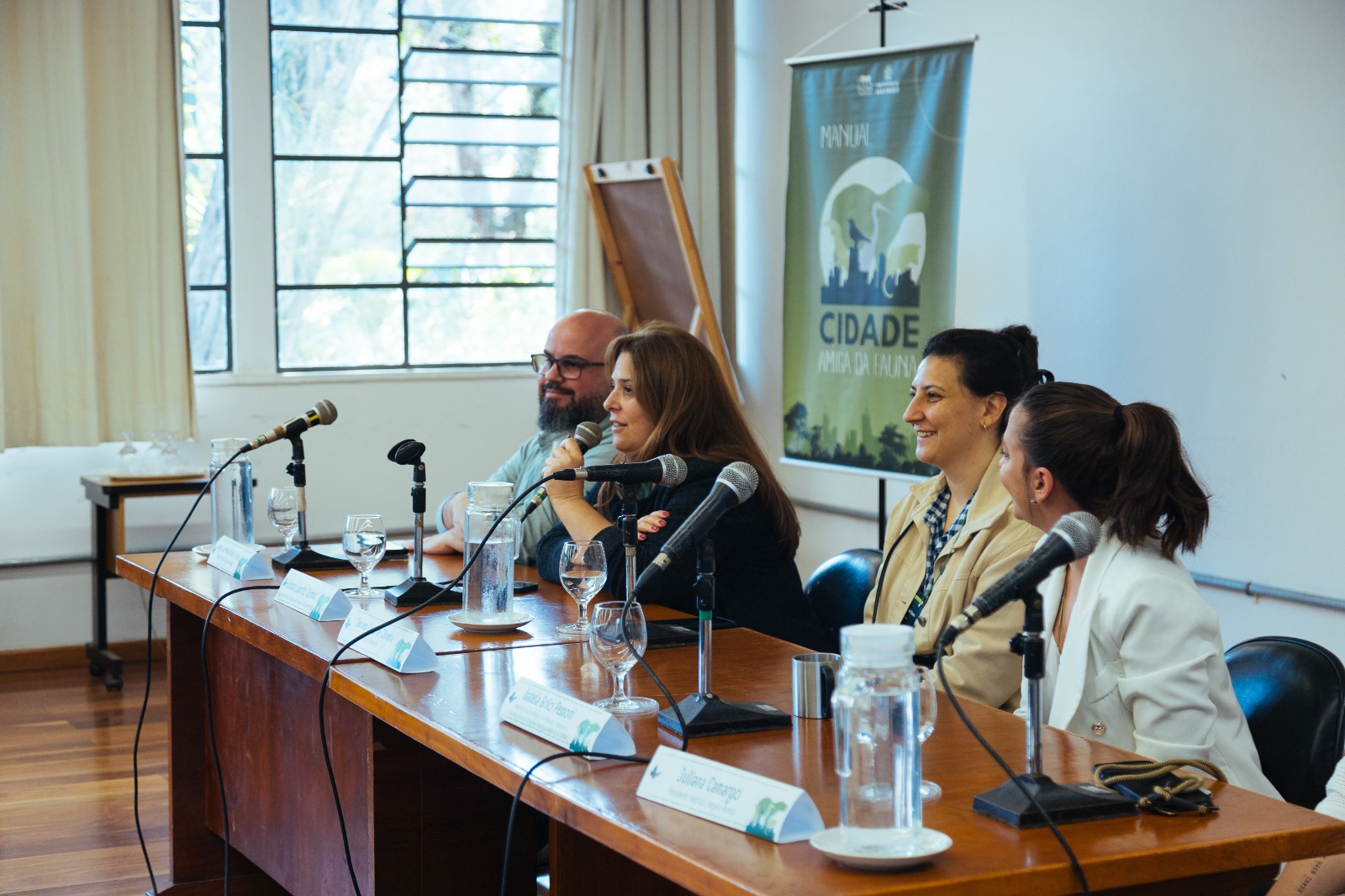 Quatro pessoas estão sentadas lado a lado atrás de uma mesa de madeira, participando de uma mesa-redonda em um ambiente interno iluminado por luz natural. Todas têm microfones à frente. Ao fundo, há um banner do “Manual Cidade Amiga da Fauna”.