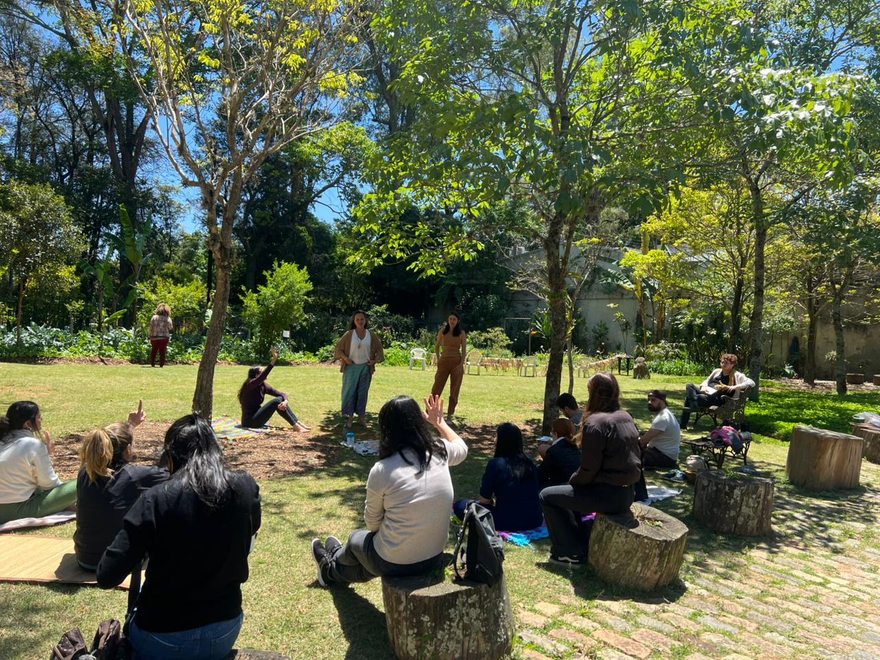Em um espaço verde arborizado, um grupo de pessoas participa de uma atividade ao ar livre. Elas estão sentadas em troncos de madeira e sobre esteiras, formando uma roda sob a sombra das árvores. Duas pessoas estão de pé, conduzindo a atividade, enquanto as demais escutam atentamente. O ambiente é ensolarado, com vegetação densa ao fundo e atmosfera tranquila de integração com a natureza.