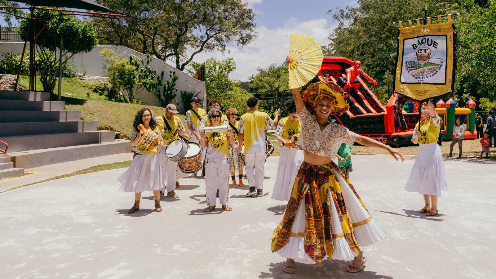 Grupo se apresenta ao ar livre com música e dança; ao centro, uma dançarina sorridente com traje colorido ergue um leque, enquanto músicos e um estandarte do grupo “Baque CT” aparecem ao fundo.