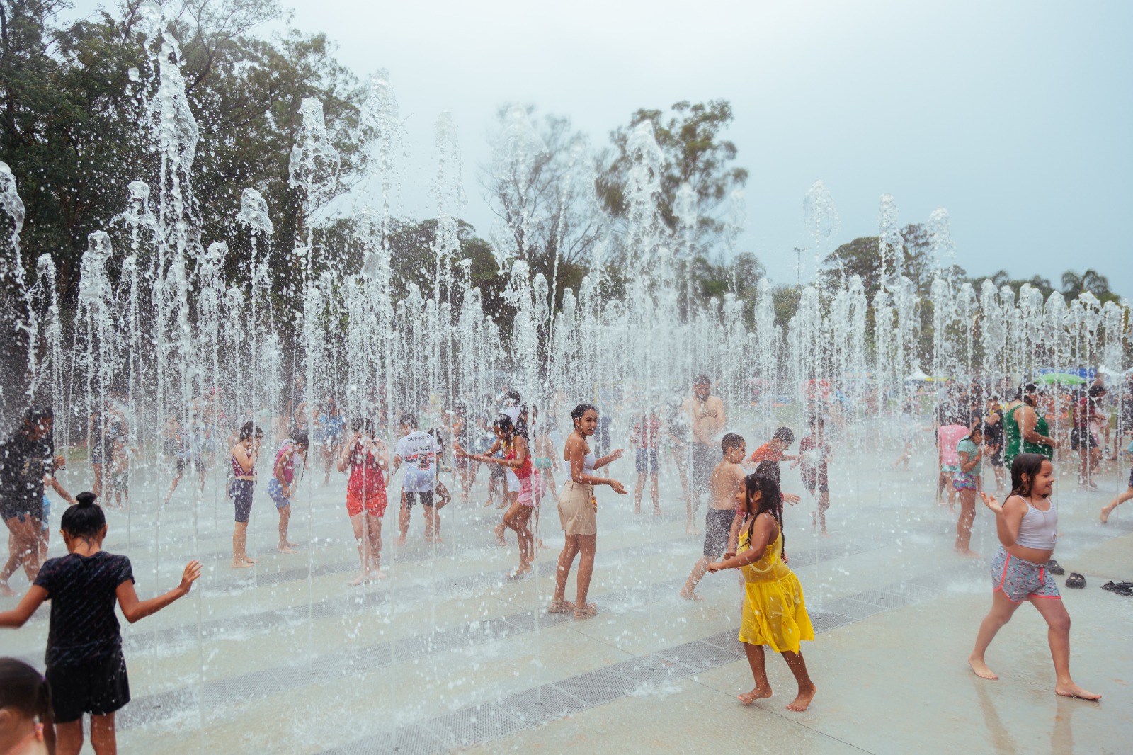 Imagem colorida de uma fonte interativa em um parque ao ar livre. Diversos jatos de água sobem do chão formando cortinas verticais. Crianças e adultos, descalços e com roupas leves de verão, brincam e caminham entre os jatos, sorrindo e se refrescando. Ao fundo, há árvores altas e o céu aparece claro e nublado, criando um clima de lazer e diversão coletiva.