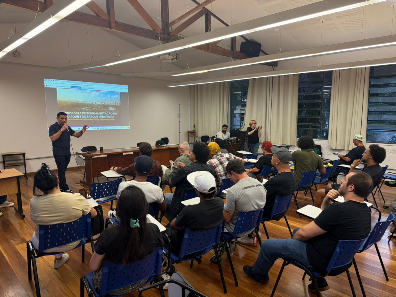 Nesta imagem, a mesma sala é vista mais de perto, destacando a área frontal onde ocorre a apresentação. Um homem em pé, vestindo camiseta escura e calça em tom neutro, segura um microfone e fala ao público, gesticulando com uma das mãos. Atrás dele, o telão branco projeta uma apresentação com predominância das cores azul e branca, com texto e imagens visíveis. As pessoas sentadas nas cadeiras azuis acompanham a fala, algumas usando bonés ou camisetas em cores como preto, cinza, verde, amarelo e vermelho. O piso de madeira marrom contrasta com as paredes claras e com as cortinas bege ao lado das janelas de vidro escuro. No canto, há equipamentos de som e projeção em tons de preto e cinza, e uma pessoa em pé registra a cena, reforçando o ambiente de reunião ou apresentação pública.