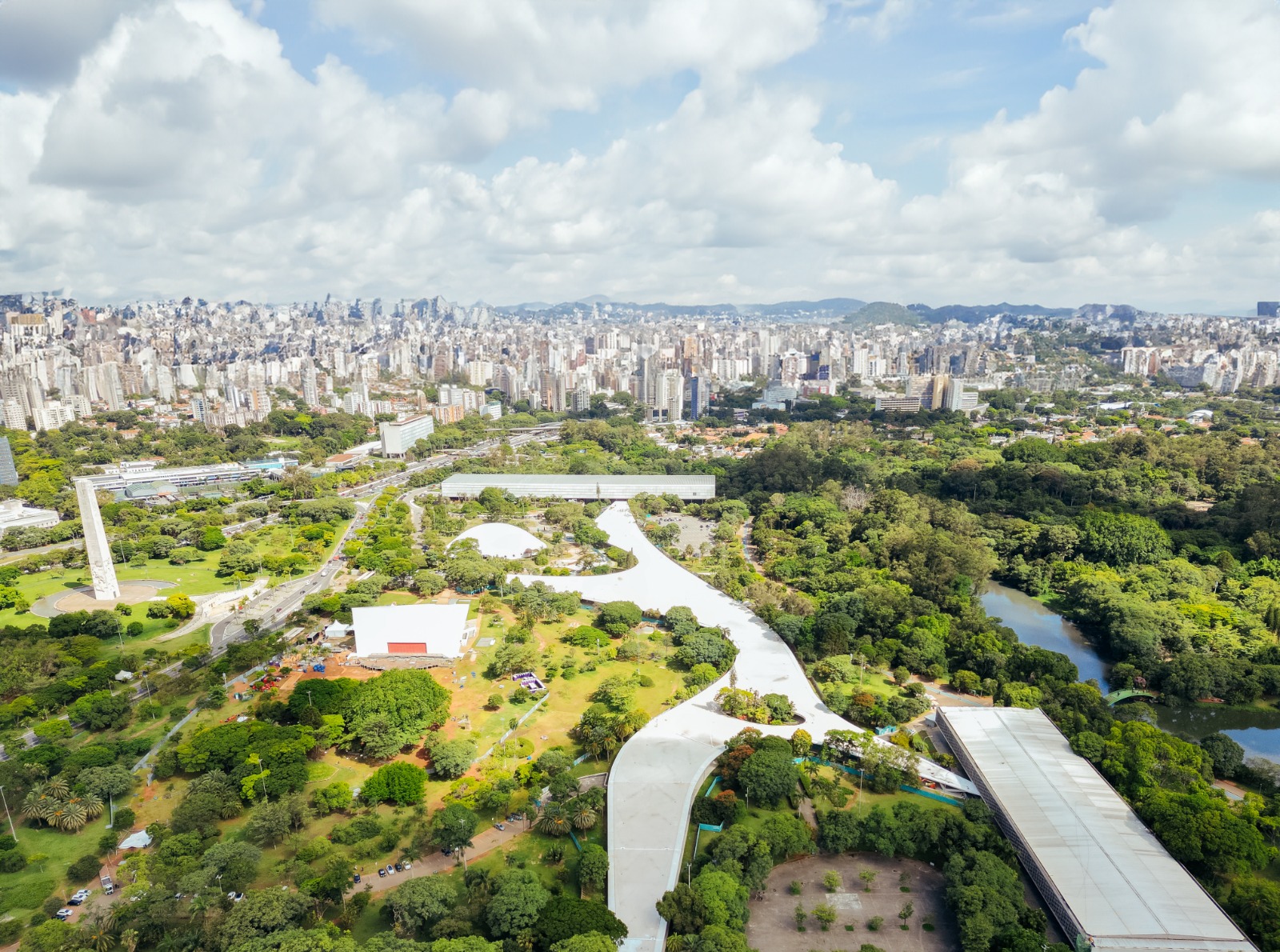 Imagem aérea panorâmica do Parque Ibirapuera, em São Paulo. Em primeiro plano, uma grande área verde com árvores densas, gramados e caminhos que se cruzam. No centro da imagem, destaca-se a Marquise do Ibirapuera, uma extensa estrutura branca, curva e contínua, com arquitetura moderna, que serpenteia pelo parque conectando diferentes espaços culturais. À direita, há um lago de formato orgânico, cercado por vegetação abundante. À esquerda, é possível ver vias internas com alguns carros e um monumento vertical claro. Espalhados pelo parque, aparecem edifícios baixos de linhas retas e telhados claros. Ao fundo, surge a paisagem urbana com muitos prédios altos e médios, criando contraste entre o verde do parque e a cidade. O céu está azul claro, com nuvens brancas, indicando um dia ensolarado.