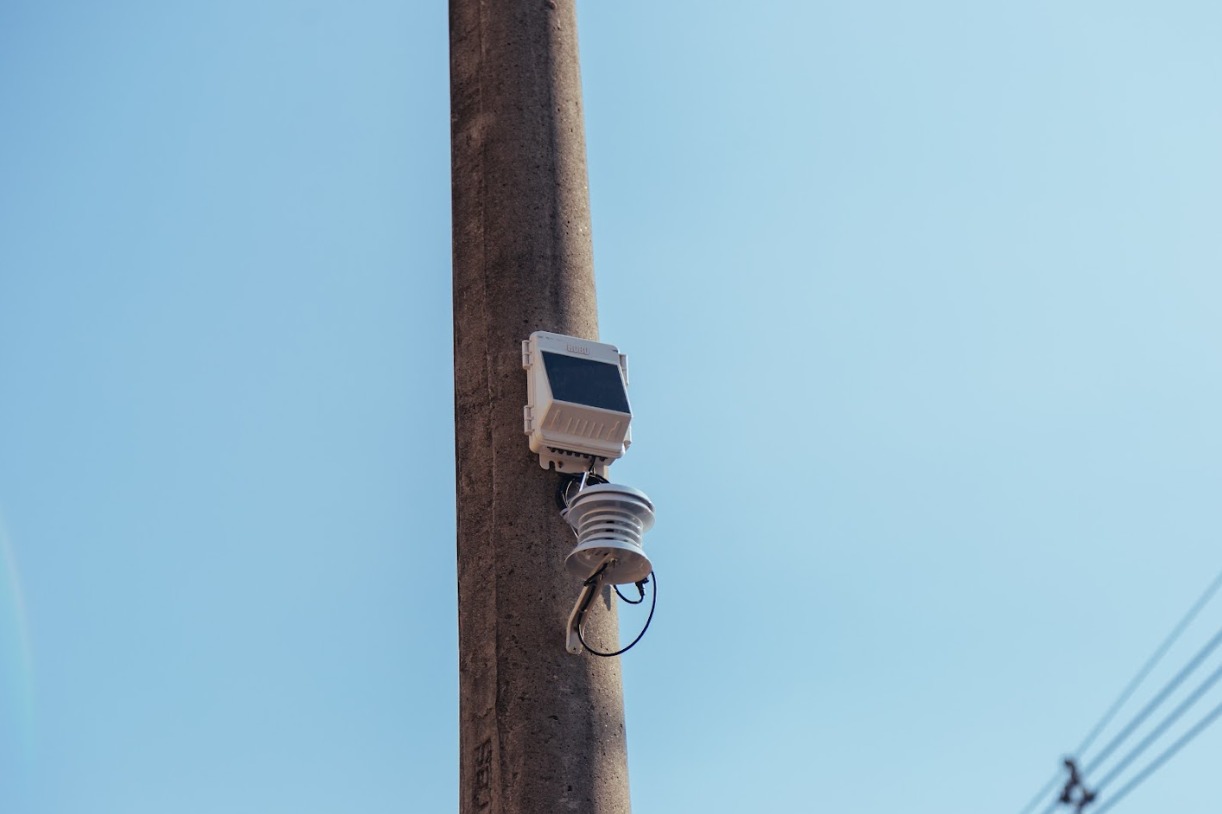 Imagem mostra um poste de concreto em tom cinza, visto de baixo para cima, ocupando a parte central da foto. Preso ao poste há um pequeno equipamento eletrônico de monitoramento, de cor branca, com um painel frontal escuro e um sensor cilíndrico logo abaixo, também em tons claros, com fios pretos visíveis. O fundo é composto por um céu azul-claro, sem nuvens, criando alto contraste com o poste. No canto inferior direito, aparecem discretamente fios elétricos escuros, parcialmente desfocados.