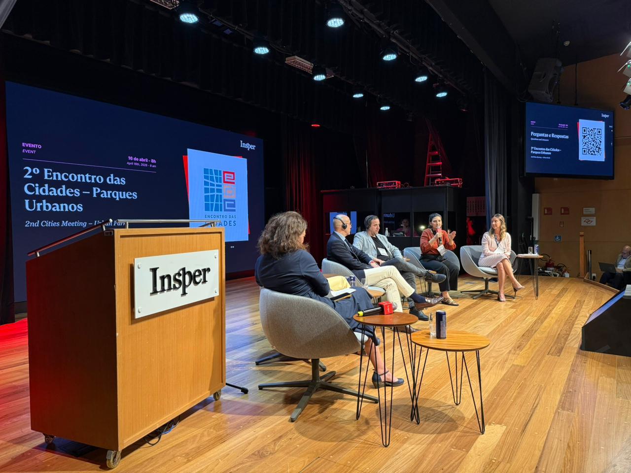 palco de auditório com painel de debate sobre parques urbanos. Cinco pessoas estão sentadas em cadeiras, uma delas falando ao microfone. Ao fundo, um telão exibe “2º Encontro das Cidades – Parques Urbanos” e o logo do Insper. À frente, há uma mesa com água e objetos de apoio. O ambiente tem iluminação de palco e plateia fora de quadro.