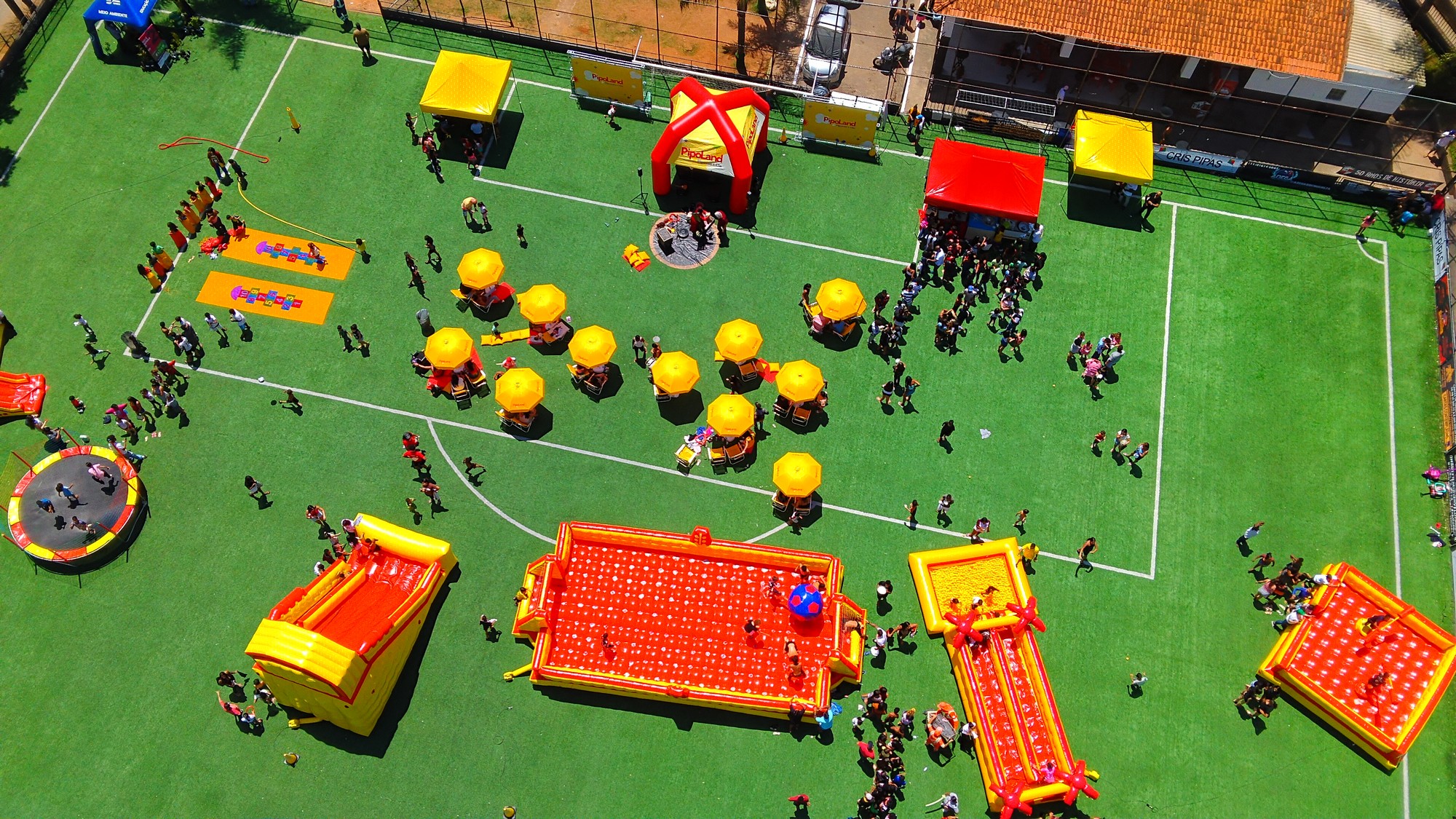 Crédito da foto: Fernando Wendling/Pipoland | #PraCegoVer: Vista aérea de um campo de futebol com grama sintética durante o evento Bairro Legal, com brinquedos infláveis vermelhos e amarelos espalhados pelo espaço, cama elástica, mesas com guarda-sóis amarelos e tendas montadas ao fundo. Crianças brincam nos infláveis enquanto adultos circulam pelo local, formando filas próximas às tendas. O dia é ensolarado e o ambiente é de lazer e convivência.