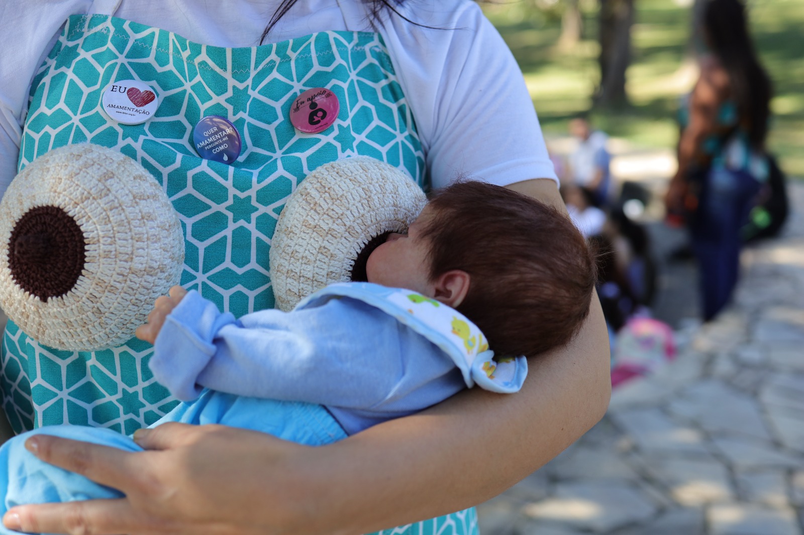 A imagem mostra uma mulher segurando um boneco que simula um bebê em posição de amamentação. Ela veste um avental azul com estampa geométrica branca, no qual estão costurados dois seios de crochê, usados como recurso educativo sobre amamentação. No avental, há também três bottons com dizeres de incentivo ao aleitamento materno: “EU ❤️ AMAMENTAÇÃO”, “QUER AMAMENTAR COMO EU?” e “Eu apoio”.  A cena ocorre ao ar livre, em ambiente arborizado, com outras pessoas visíveis ao fundo, desfocadas. A imagem retrata uma ação de sensibilização e promoção da amamentação, possivelmente durante uma oficina ou evento ligado ao Agosto Dourado.         