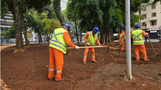 A foto mostra um grupo de trabalhadores da manutenção ou jardinagem preparando o solo em uma área verde urbana, possivelmente uma praça.  Cerca de quatro homens estão de frente ou perfil, vestindo uniformes de segurança laranja e coletes refletivos amarelo-limão, além de capacetes de segurança azuis. Eles estão usando enxadas para nivelar e afofar a terra vermelha e argilosa do canteiro.  Ao fundo, há grandes árvores de folhagem densa, proporcionando sombra. Mais ao fundo, à esquerda e à direita, há o ambiente urbano com o trânsito de uma rua e prédios de vários andares, indicando que a cena ocorre em uma área central da cidade. Um poste de iluminação cinza está visível à direita.