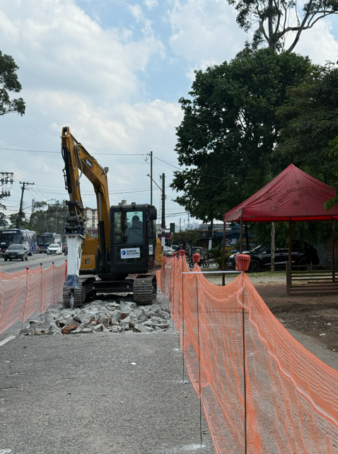 A imagem mostra uma obra em uma via pública durante o dia. Ao centro, uma escavadeira amarela com detalhes em preto e cabine fechada está posicionada de frente, equipada com um martelete hidráulico que repousa sobre um trecho de asfalto quebrado e pedaços de concreto. A área de trabalho é delimitada por uma tela de proteção plástica na cor laranja, esticada em ambos os lados da máquina. À direita, em primeiro plano, a cerca laranja segue em perspectiva, e ao fundo há uma tenda vermelha montada sob árvores verdes. À esquerda, observa-se o fluxo de uma avenida com ônibus azul e carros brancos em movimento. O céu está parcialmente nublado, e na porta da cabine da escavadeira há um logotipo com o nome "Ramon Aguilera"