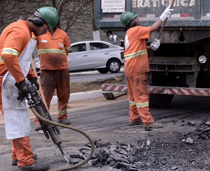 A imagem mostra uma equipe de três trabalhadores da construção civil vestindo uniformes laranjas e capacetes verdes. Eles estão em uma rua, com um dos trabalhadores operando uma britadeira para quebrar uma pilha de asfalto. Um outro trabalhador, usando um avental branco, observa o trabalho. Um terceiro trabalhador está posicionado perto de um grande caminhão verde, usando uma pá para mover os detritos. No fundo, um carro branco está estacionado.