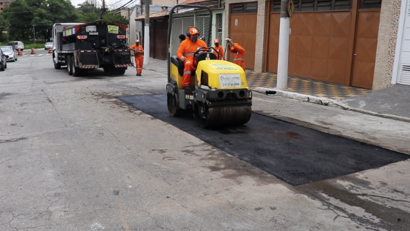 A imagem mostra uma rua em um bairro residencial. No centro, um trabalhador da construção civil com uniforme e capacete laranja opera um rolo compressor amarelo, compactando uma nova seção de asfalto. Um segundo trabalhador, também de uniforme laranja, está perto do rolo, ajudando no trabalho. No fundo, um grande caminhão preto está estacionado, com mais dois trabalhadores por perto. A rua é ladeada por casas e edifícios de apartamentos em ambos os lados. O céu está encoberto.
