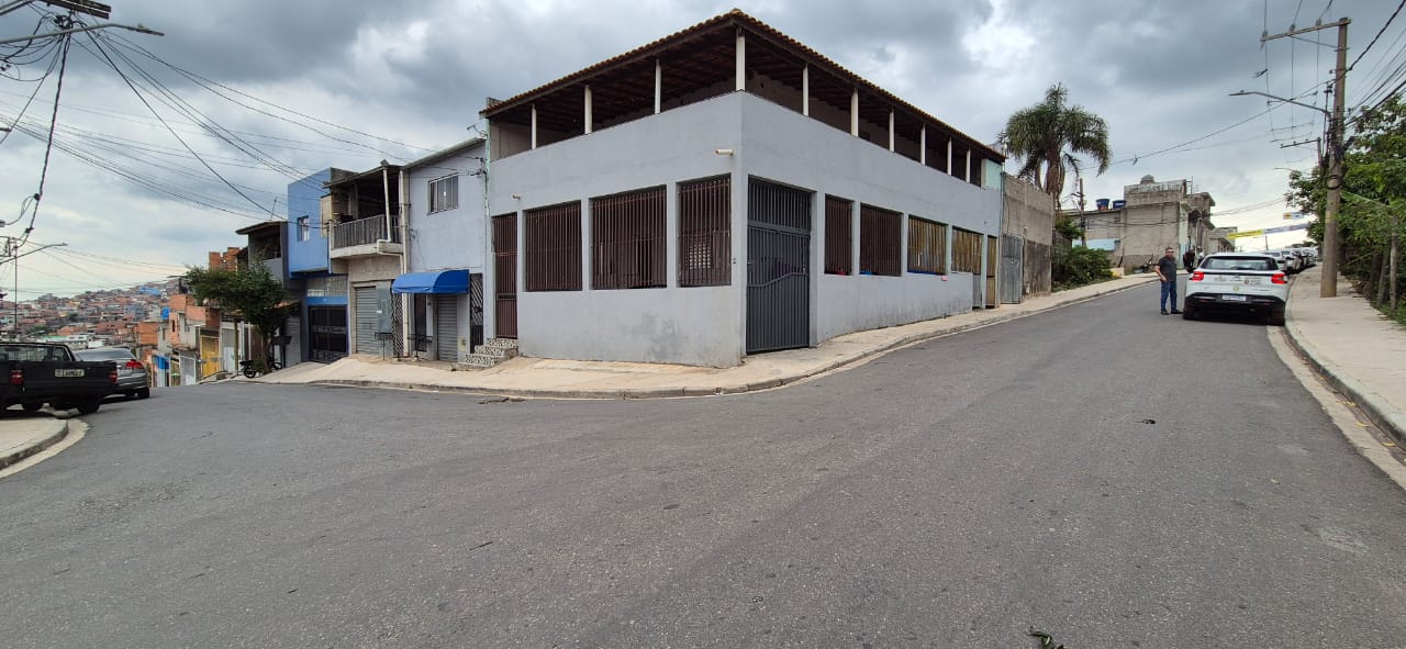 A imagem é um registro de uma esquina em um bairro de periferia, sob um céu nublado. No centro da foto, destaca-se um prédio de esquina de dois andares, pintado de cinza claro. O andar térreo possui grades e um portão de ferro, e o andar superior é uma ampla varanda. A rua principal no primeiro plano é asfaltada e se curva para a direita, onde um carro SUV branco está estacionado e um homem de camisa clara está em pé. À esquerda, a rua desce abruptamente, revelando casas coloridas e, ao longe, uma vasta paisagem urbana densamente construída, característica de uma comunidade em área de encosta. Fios e cabos elétricos cruzam o topo da cena.