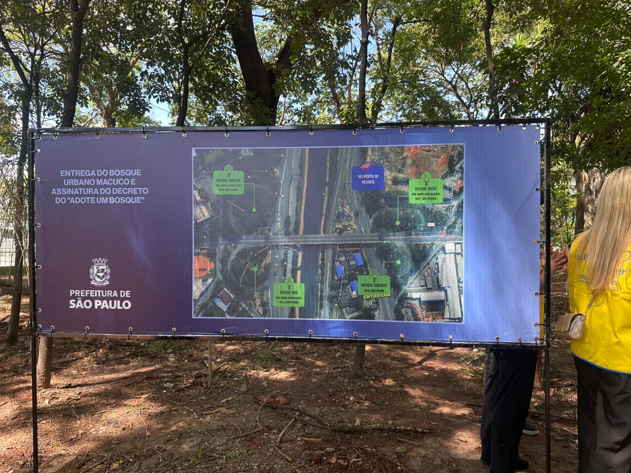 A foto mostra um grande painel informativo de lona azul montado em uma estrutura metálica no meio de uma área arborizada. No lado esquerdo do painel, lê-se o texto em branco: "ENTREGA DO BOSQUE URBANO MACUCO E ASSINATURA DO DECRETO DO 'ADOTE UM BOSQUE'", acompanhado pelo logotipo da Prefeitura de São Paulo. Ao centro, há um mapa aéreo da região destacando diferentes áreas verdes com etiquetas coloridas: o Bosque Siriema, o Bosque Boi e o Bosque Irerê são marcados como "em implantação", enquanto o Bosque Macuco aparece com a etiqueta verde "entregue". O painel está instalado sobre um solo de terra e folhas secas, cercado por árvores cujas sombras são projetadas no chão. À direita, parte das costas de uma pessoa com colete amarelo e cabelos loiros aparece na borda do enquadramento.