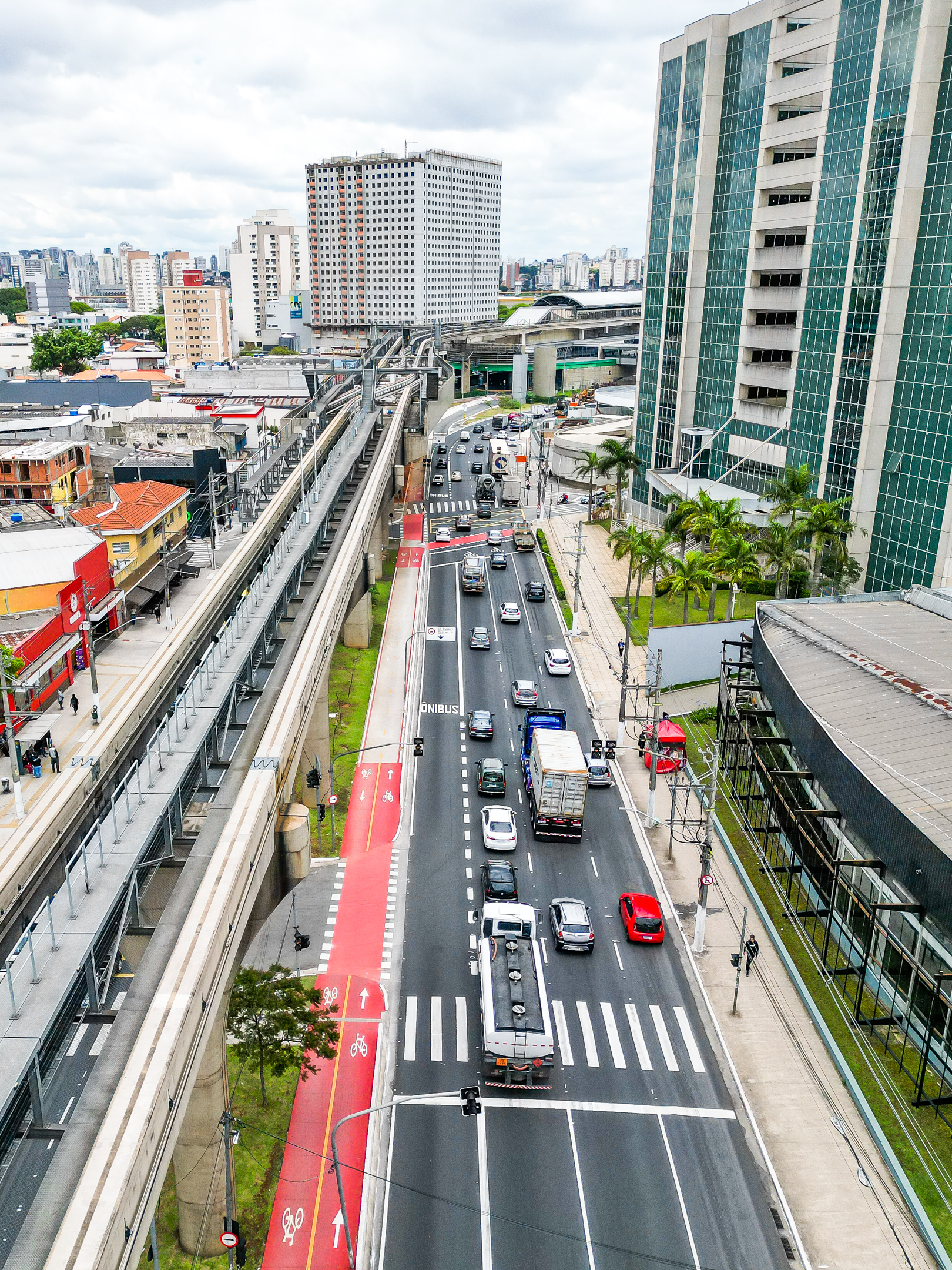 A imagem é uma fotografia aérea vertical, em plano aberto, de uma grande avenida urbana. À esquerda, estendem-se os trilhos elevados de concreto de um sistema de monotrilho, que correm paralelos à via. Logo ao lado dos pilares do monotrilho, há uma ciclovia de mão dupla pintada em vermelho vibrante. A avenida, ao centro, apresenta asfalto escuro recém-recapeado com sinalização branca nítida, onde transitam carros, um caminhão baú e um caminhão-tanque. À direita da avenida, observa-se uma calçada larga com palmeiras em frente a um imponente edifício comercial moderno com fachada de vidro esverdeado. Ao fundo, a paisagem é composta por diversos prédios residenciais e comerciais sob um céu nublado.