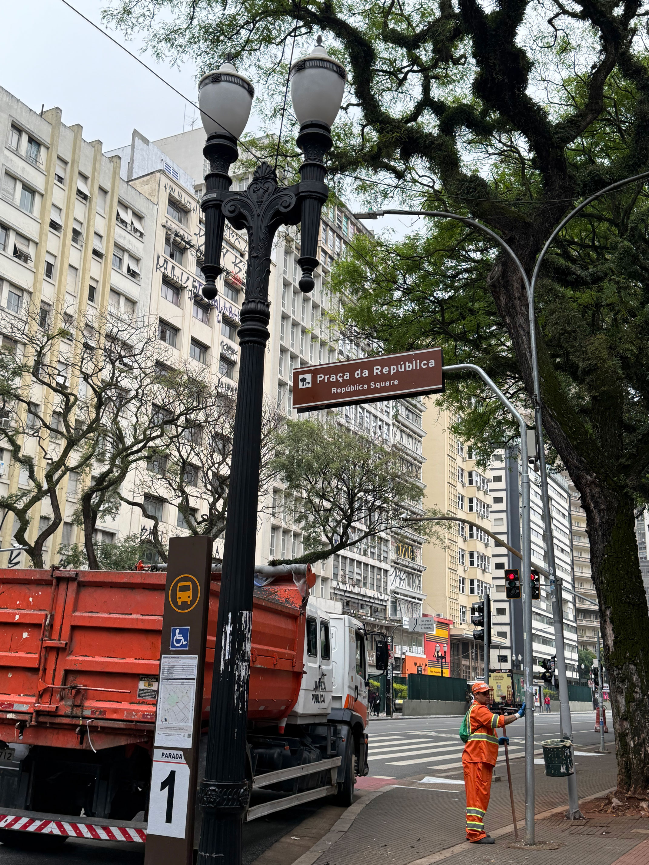 Fotografia em um centro urbano. Em primeiro plano, no centro, há um poste de luz antigo, alto e de ferro preto, com duas luminárias brancas. À esquerda do poste, estacionado na rua, está a parte traseira de um caminhão de lixo grande, com caçamba de cor laranja vibrante. Há também uma placa marrom indicando um ponto de ônibus e acessibilidade. Mais ao fundo e à direita, um trabalhador vestido com uniforme laranja e capacete branco, segurando um instrumento, parece estar limpando a calçada ou a rua, próximo a um semáforo vermelho. Acima do trabalhador, uma placa de rua marrom diz "Praça da República". Ao fundo, prédios altos e antigos com muitas janelas se estendem para a esquerda e para a direita, e grandes árvores frondosas ladeiam a rua. O céu está claro, mas nublado.